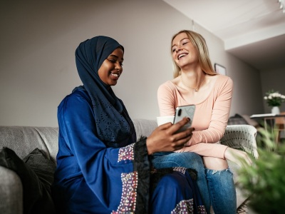 En kvinne med hijab og en kvinne uten hijab sitter i en sofa. De er venninner og ler av noe de ser på mobilen. Foto