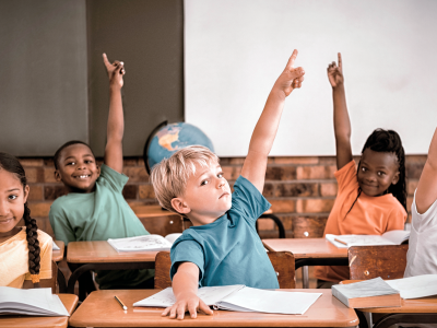 Seks barn sitter i klasserom bak skrivebord. Alle rekker ei hånd opp i lufta. Foto.