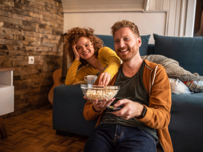 En mann sitter på gulvet med popcorn, mens en kvinne ligger på sofaen rett bak ham. De ser på TV. Foto