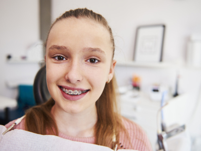 En ung jente med tannregulering sitter i tannlegestolen. Hun smiler mot kamera. Foto