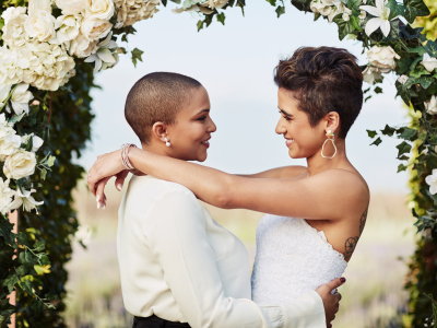 To kvinner står under en plomsterport og ser hverandre inn i øynene. En har på seg brudekjole og en har på seg hvit skjorte. De smiler og er glade. Foto