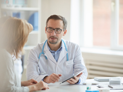 En mannlig lege med stetoskop rundt halsen snakker med en kvinnelig pasient. Foto