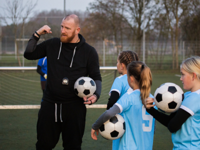 En mann og tre jenter står med hver sin fotball på en fotballbane. Mannen gir instruksjoner. Han er faren til en av jentene og treneren til jentelaget. Foto