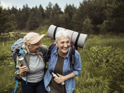 To kvinner i 70-årene er på tur i skogen. De ler og er gode venner. Begge har på seg sekker med turutstyr og holder vannflasker i henda. Bildet illustrerer et aktivt pensjonistliv. Foto