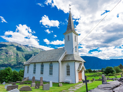 En hvit kirke. Utenfor er det en kirkegård. Bak kirken kan vi se fjell og en blå himmel med noen skyer. Foto