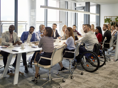 Mange ansatte sitter rundt et bord. De er på jobbmøte. De ansatte er kvinner og menn av forskjellige hudfarger, og en person sitter i rullestol. Bildet illustrerer yrkesdeltakelse. Foto