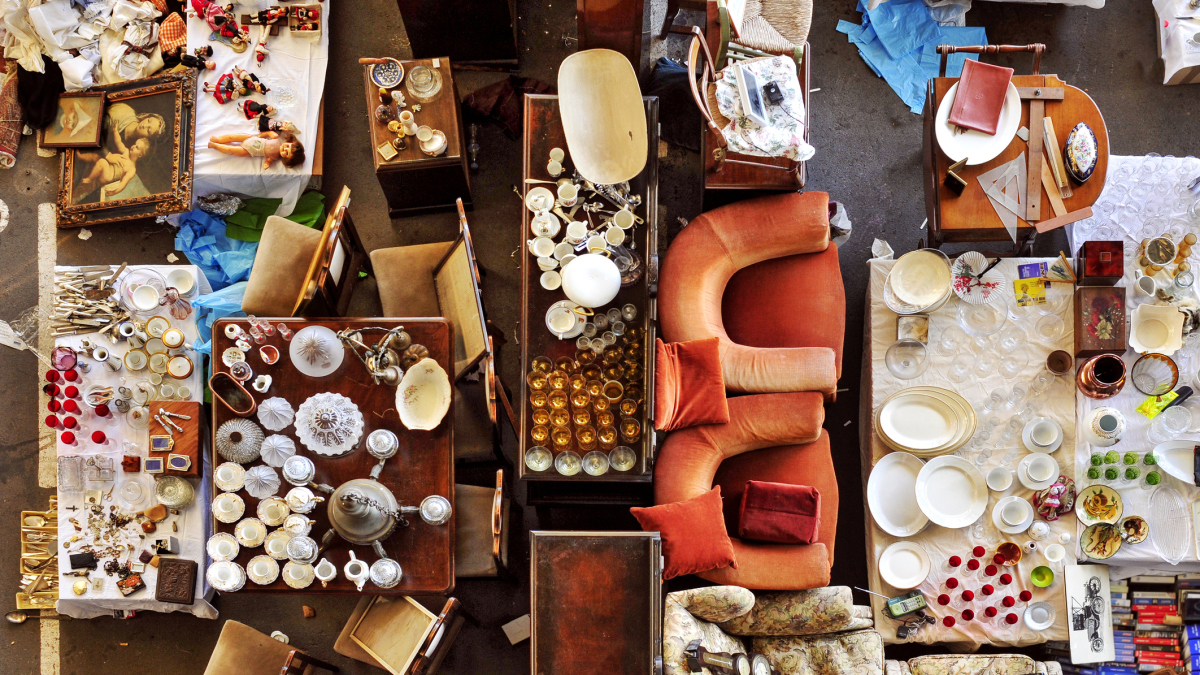 Et bilde tatt ovenfra. Vi ser en sofa og sofastoler. På bord er det plassert tallerkener, kopper og kar. Bildet er fra et loppemarked.