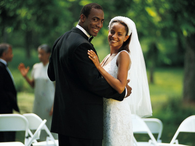 En mann og en kvinne smiler mot kamera. Han har på seg dress, og hun har på seg brudekjole. De er glade. Foto