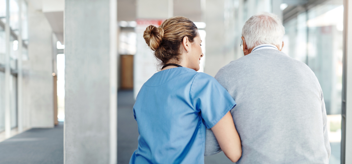 En ung sykepleier støtter en eldre mann når han går. Bildet er tatt bakfra, og man kan se at de snakker sammen. Foto