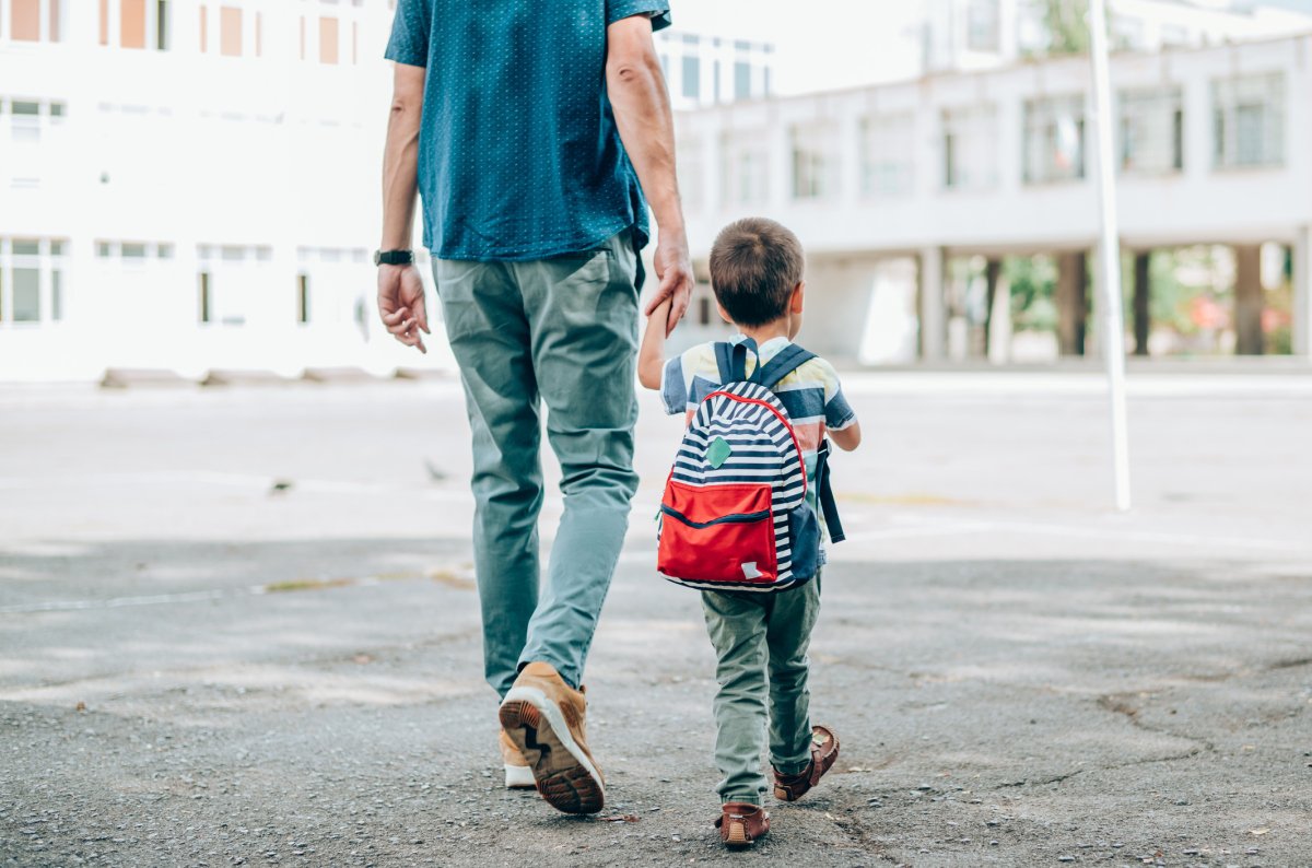 En voksen far holder sin lille sønn i hånden. Sønnen har på seg en ryggsekk. De er på vei til skolen. Foto