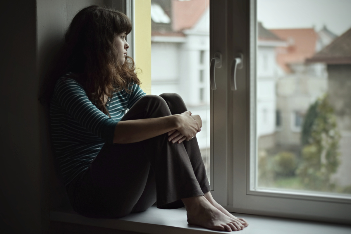 En ung kvinne sitter i vinduskarmen. Hun er trist og betenkt mens hun ser ut av vinduet. Bildet illustrerer en persons manglende frihet når hun blir utsatt for negativ sosial kontroll. Foto